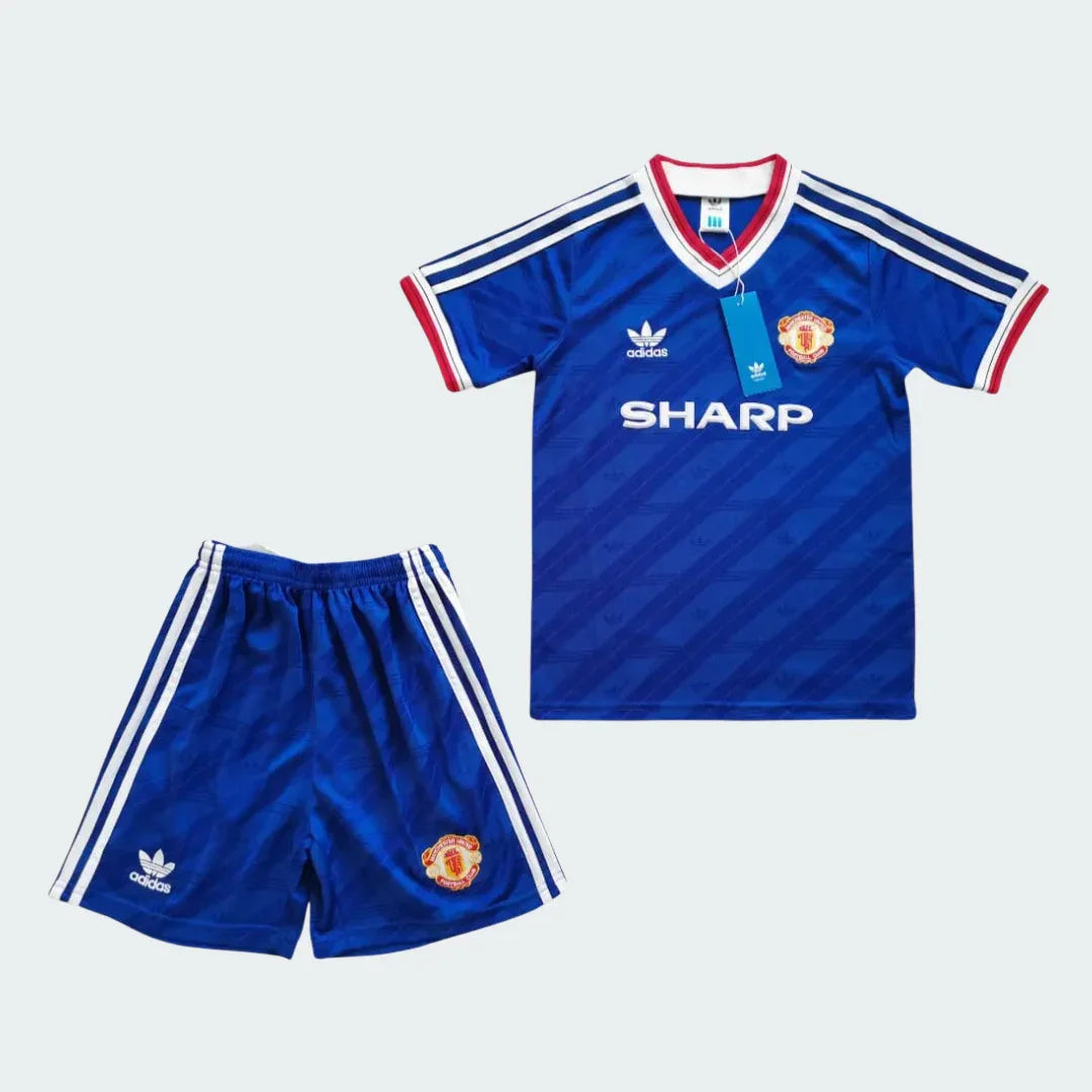 Kit infantil azul do Manchester United 1986, com camisa e shorts. Inclui escudo do time, logo Adidas, patrocinador Sharp e listras brancas.