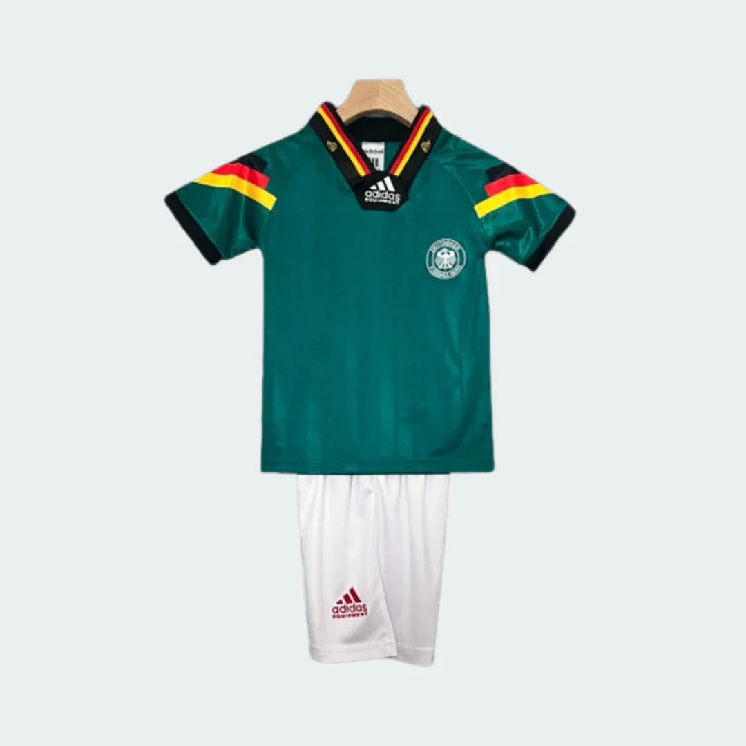 Kit infantil retrô da Alemania II 1992 completo. Camisa verde escura com gola V preta, listras pretas, vermelhas e amarelas nas mangas. Escudo da Alemania no peito. Shorts brancos com logo Adidas.