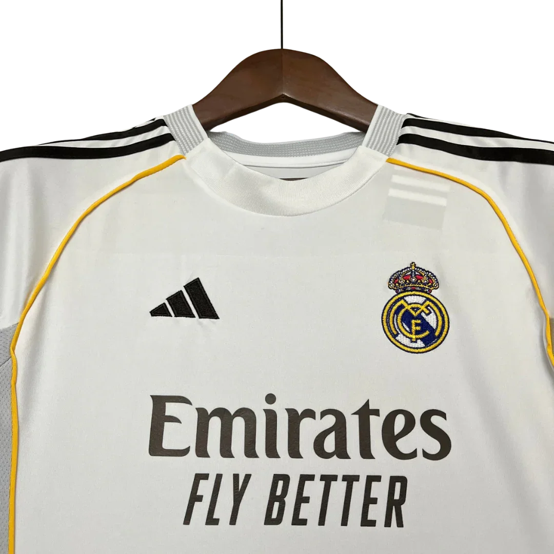 Close-up de camisa branca do Real Madrid em cabide, com escudo do time, logo Adidas e patrocinador Emirates. Detalhes amarelos e listras pretas nos ombros.