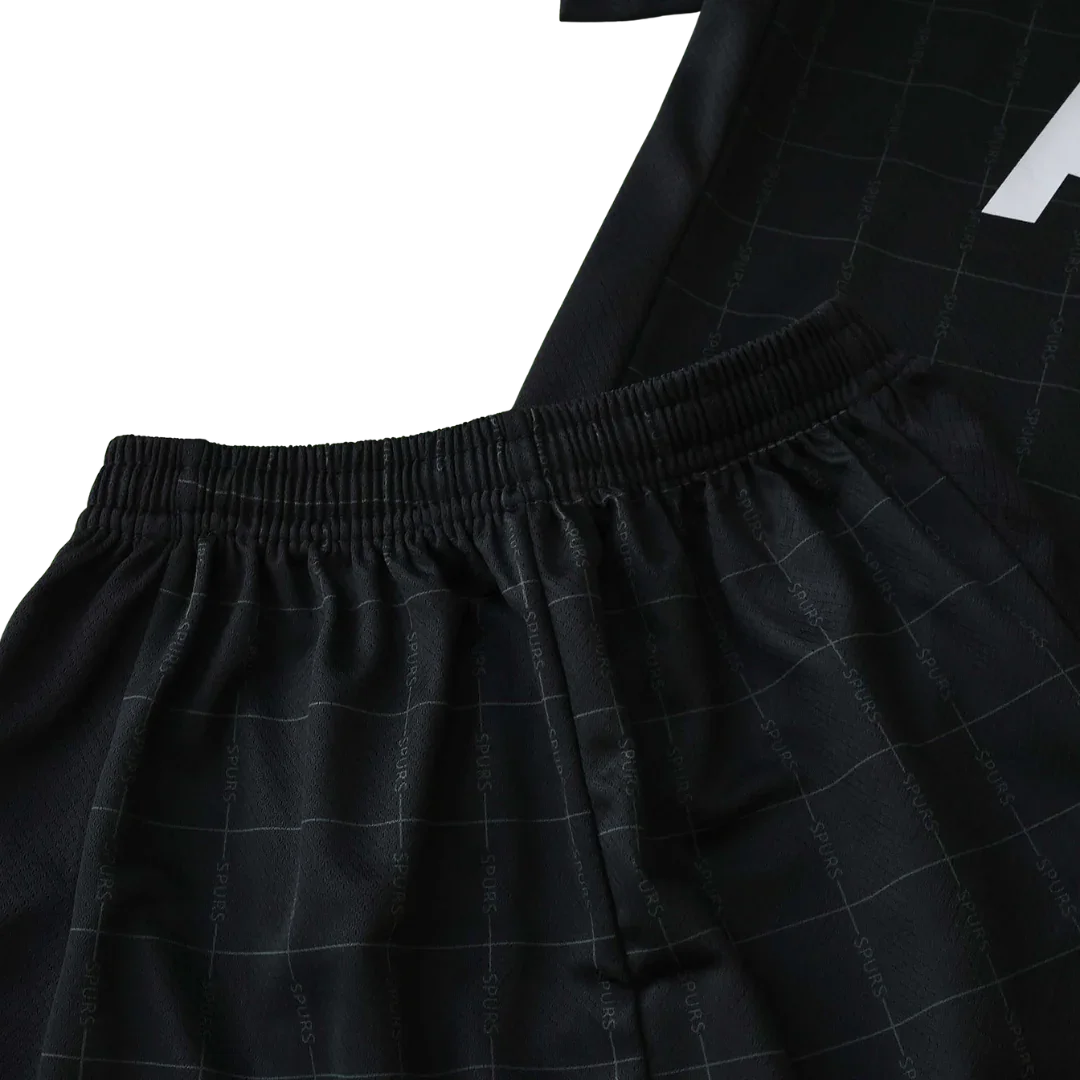 Detalhe de um kit infantil preto, mostrando shorts e parte de uma camisa. O tecido possui padrão quadriculado e texto repetido. Cós elástico e canelado.