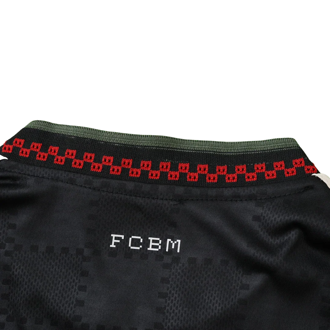 Vista traseira de camisa de futebol infantil preta com textura. Gola verde e faixa vermelha estampada. Letras FCBM brancas atrás.