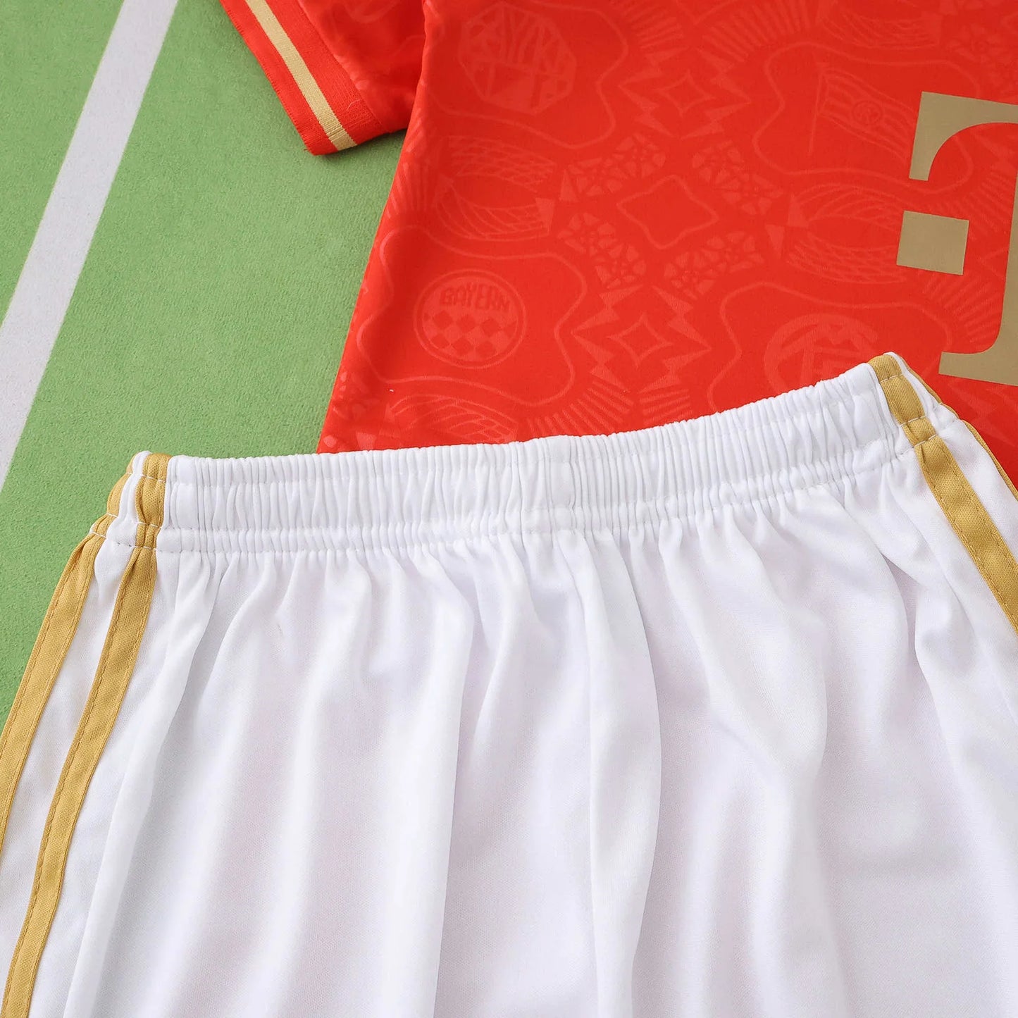 Kit de futebol infantil com camisa vermelha (logo T dourado e escudo em relevo) e shorts brancos com listras douradas.