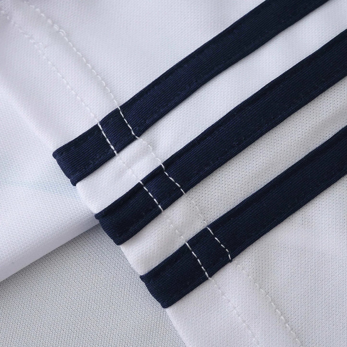 Detalhe aproximado de tecido de camisa branca com três listras diagonais em azul marinho. Costuras brancas nas bordas das listras são visíveis.