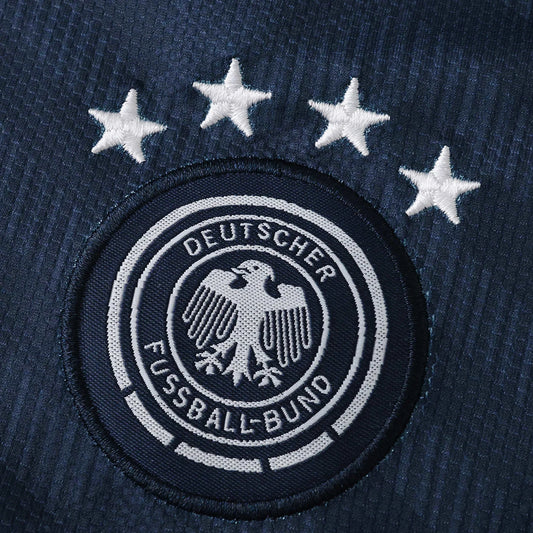 Detalhe do escudo bordado da seleção da Alemanha, com o brasão da DFB e a frase 'DEUTSCHER FUSSBALL-BUND'. Quatro estrelas brancas de campeão mundial estão acima do escudo, tudo em tecido azul marinho.