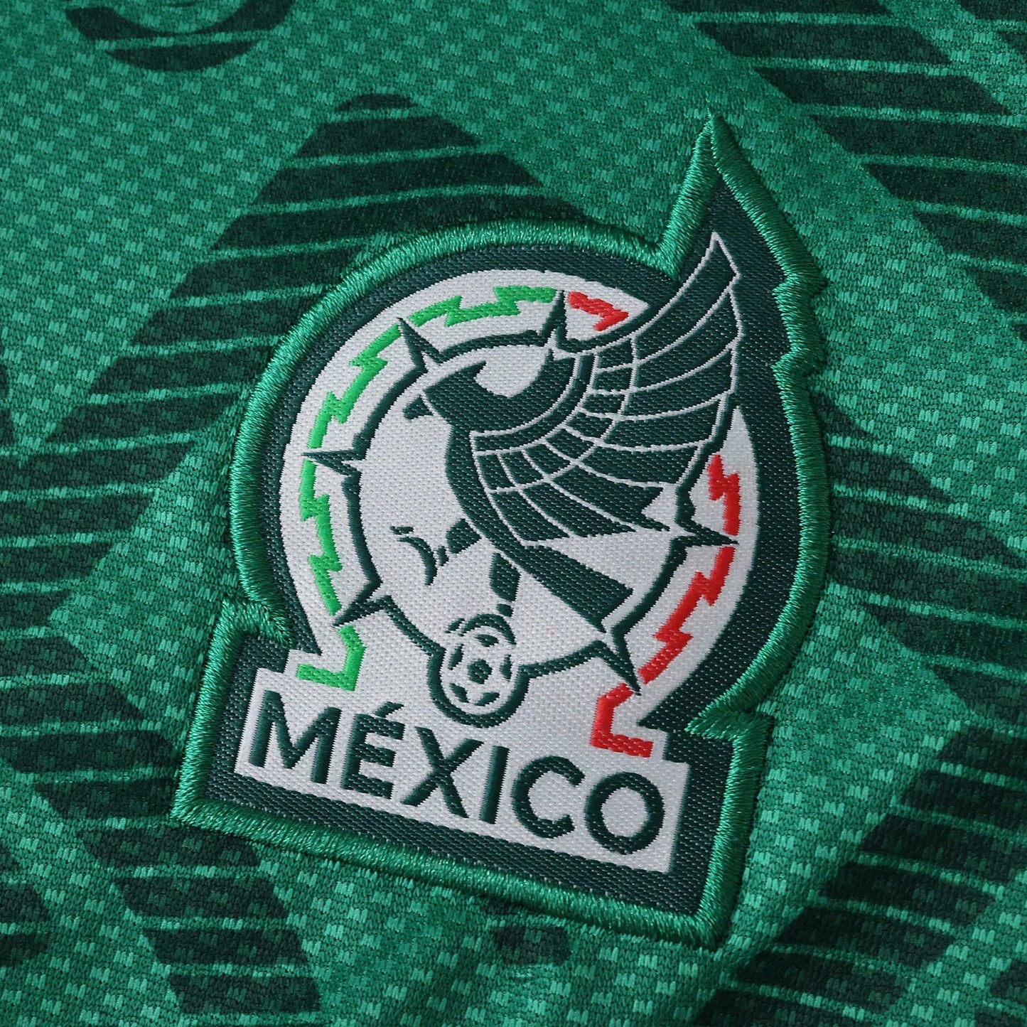 Escudo bordado da seleção do México em camisa verde escura com padrão. Águia, bola de futebol, cores verde, branco e vermelho.