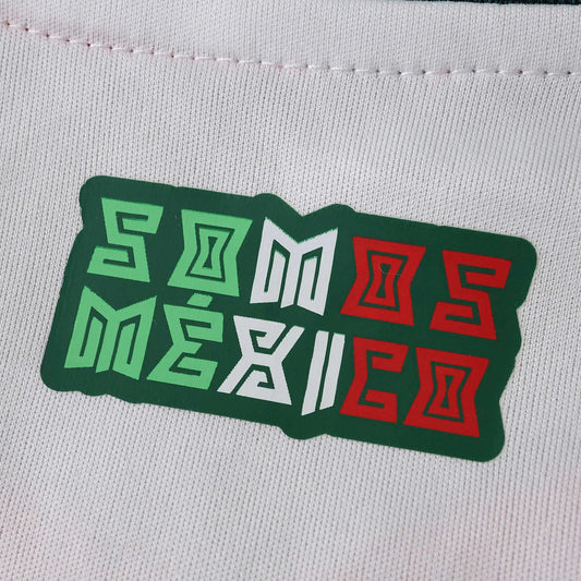 Close-up de tecido de camiseta branca com emblema verde 'SOMOS MÉXICO'. O texto combina letras verdes, brancas e vermelhas.