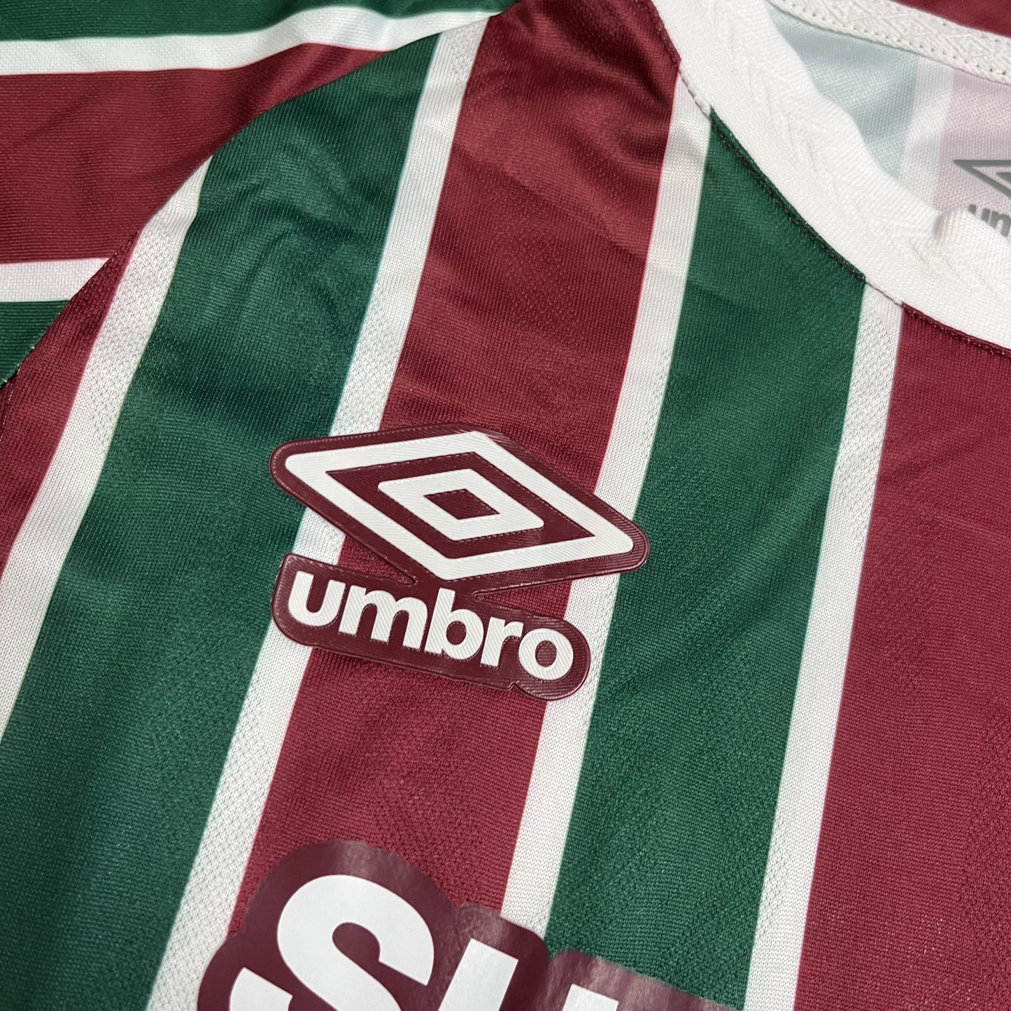 Close do logotipo da Umbro bordado em branco com contorno grená, posicionado sobre uma das listras verdes da camisa.