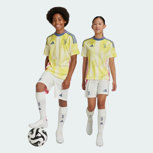 Menino e menina sorrindo com kit Juventus II 24/25. Camisa amarela e branca com gola azul, shorts e meias creme. Emblemas Juventus e Adidas visíveis.