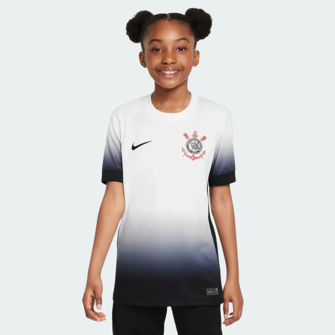 Menina sorridente vestindo camisa de futebol infantil do Timão branca com degradê para preto, logo Nike e escudo do clube.