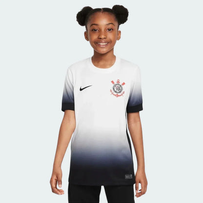 Menina sorridente vestindo camisa de futebol infantil do Timão branca com degradê para preto, logo Nike e escudo do clube.