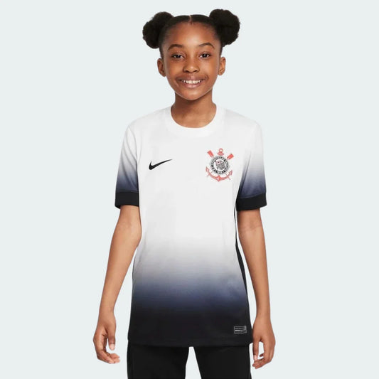 Menina sorridente vestindo camisa de futebol infantil do Timão branca com degradê para preto, logo Nike e escudo do clube.