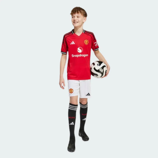 Menino sorridente em pé, vestindo o Kit Infantil Manchester United I com camisa vermelha, shorts brancos e meias pretas, segurando uma bola de futebol.