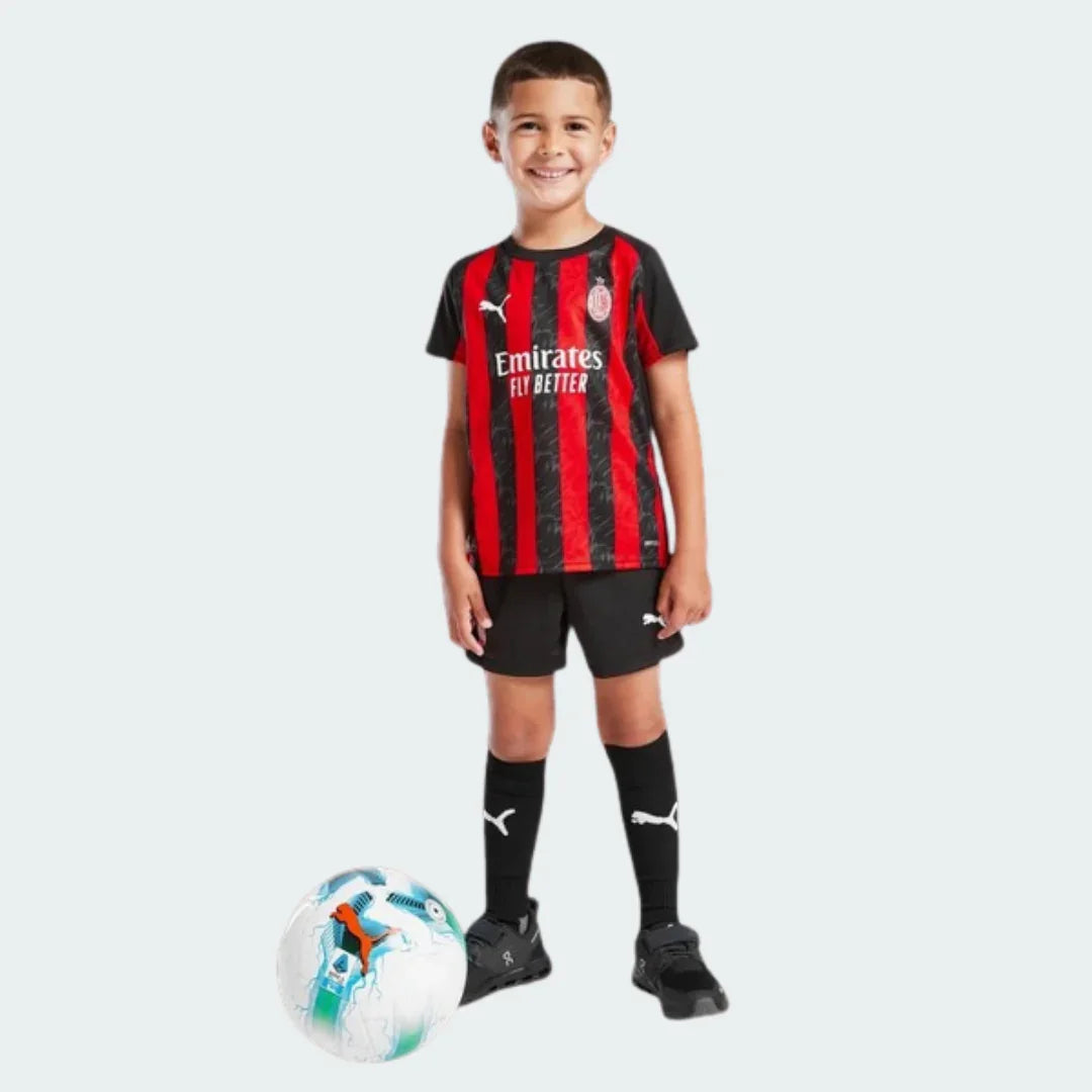 Menino sorridente veste kit de futebol infantil do Milan com camisa listrada preta e vermelha, shorts pretos e meias pretas. Bola de futebol no chão.