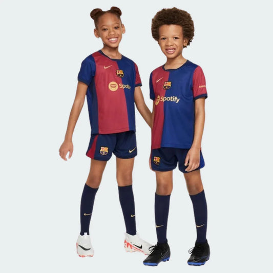 Duas crianças sorrindo, uma menina e um menino, vestindo o kit infantil do Barcelona. A camisa tem faixas verticais azul e grená, shorts e meias azuis.