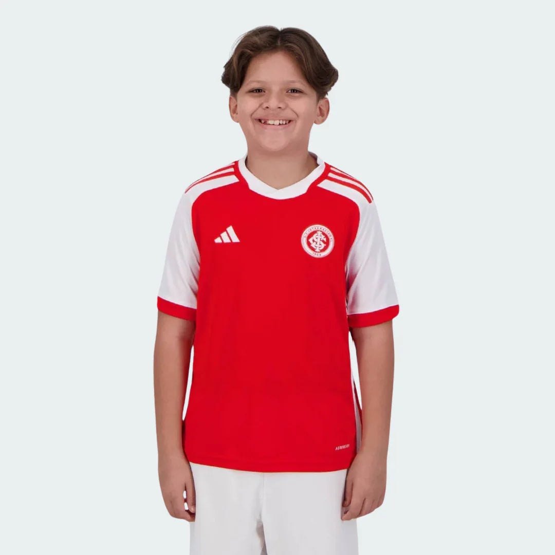 Menino sorrindo veste kit infantil Colorado I 24/25. Camisa vermelha e branca com escudo do Internacional e logo Adidas. Shorts brancos.