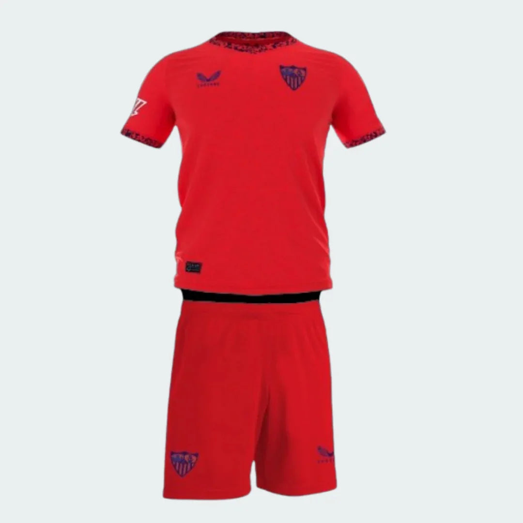 Kit de futebol infantil Sevilla 24/25. Camisa e shorts vermelhos com escudos Sevilla e logos Castore em azul escuro.