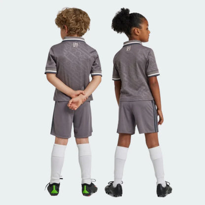 Duas crianças de costas, um menino e uma menina, vestindo kits de futebol cinzas. Camisas com logo 'RM' na gola, shorts e meias brancas.
