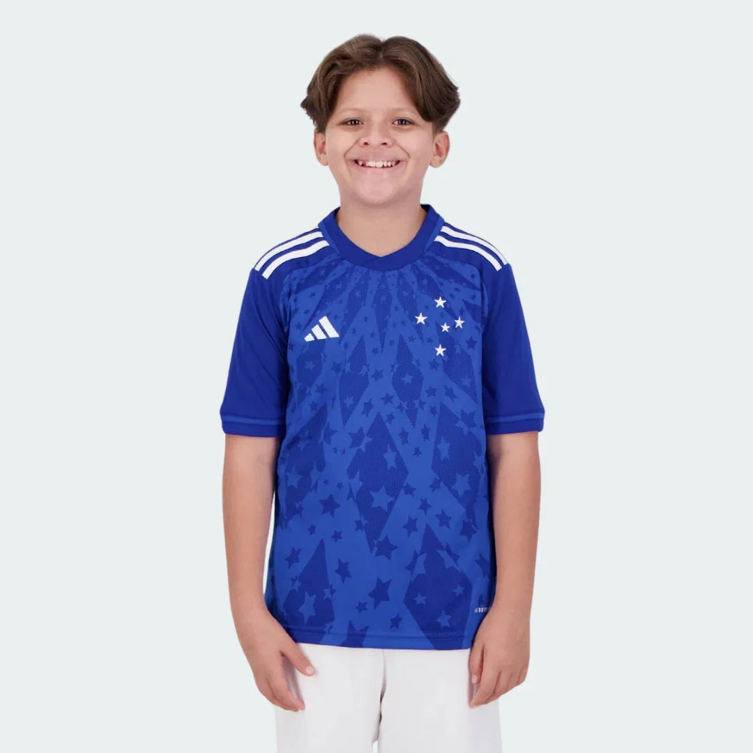 Criança feliz vestindo kit de futebol infantil Cabuloso I 24/25 azul com estrelas brancas e logo Adidas em fundo branco.