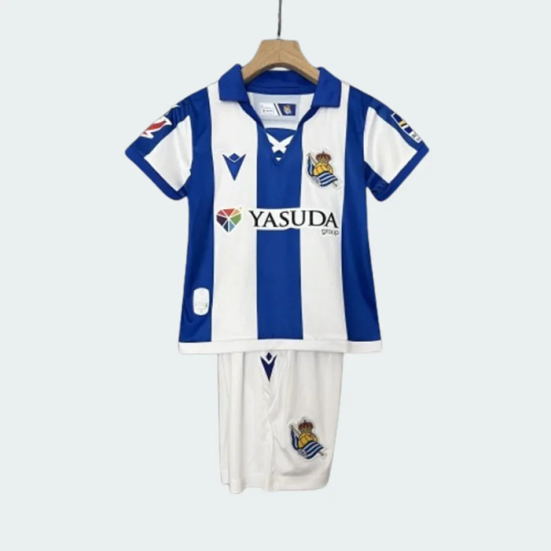 Kit infantil Real Sociedad I 24/25 com camisa listrada azul e branca, gola polo, escudo do time e shorts brancos com escudo.