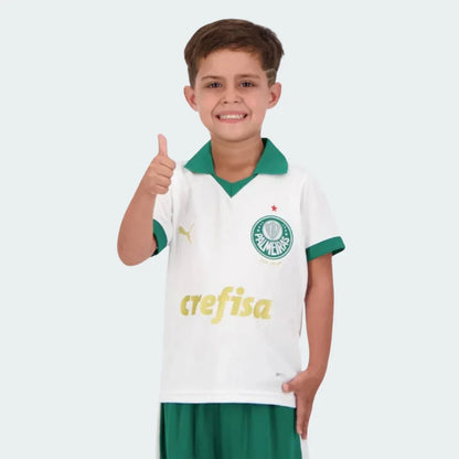 Menino sorridente em kit infantil Verdão branco com gola verde, distintivo Palmeiras, logo Puma e patrocínio Crefisa dourado, fazendo 'legal'.