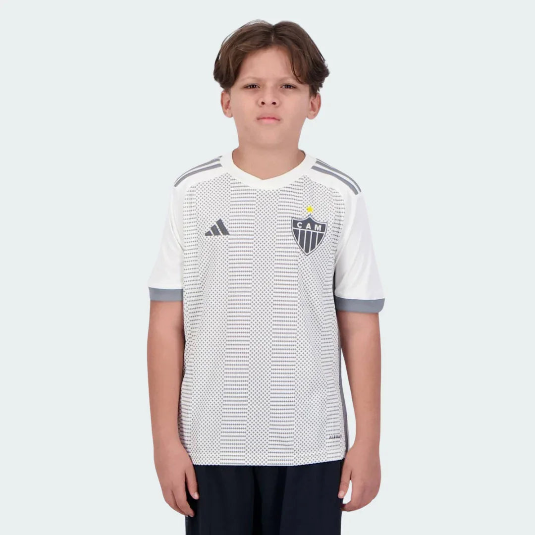 Menino usa camisa infantil branca do Galo 24/25 com padrão pontilhado cinza, escudo CAM, logo Adidas e shorts escuros.