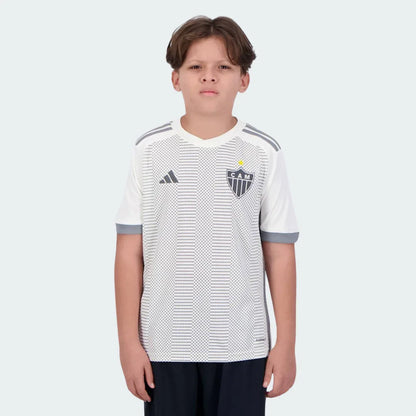 Menino usa camisa infantil branca do Galo 24/25 com padrão pontilhado cinza, escudo CAM, logo Adidas e shorts escuros.