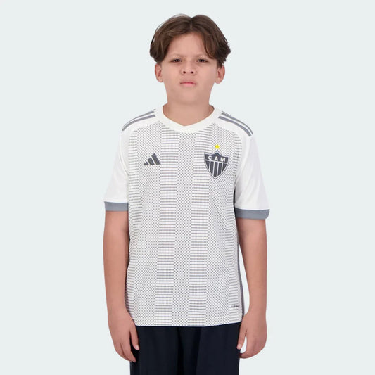 Menino usa camisa infantil branca do Galo 24/25 com padrão pontilhado cinza, escudo CAM, logo Adidas e shorts escuros.