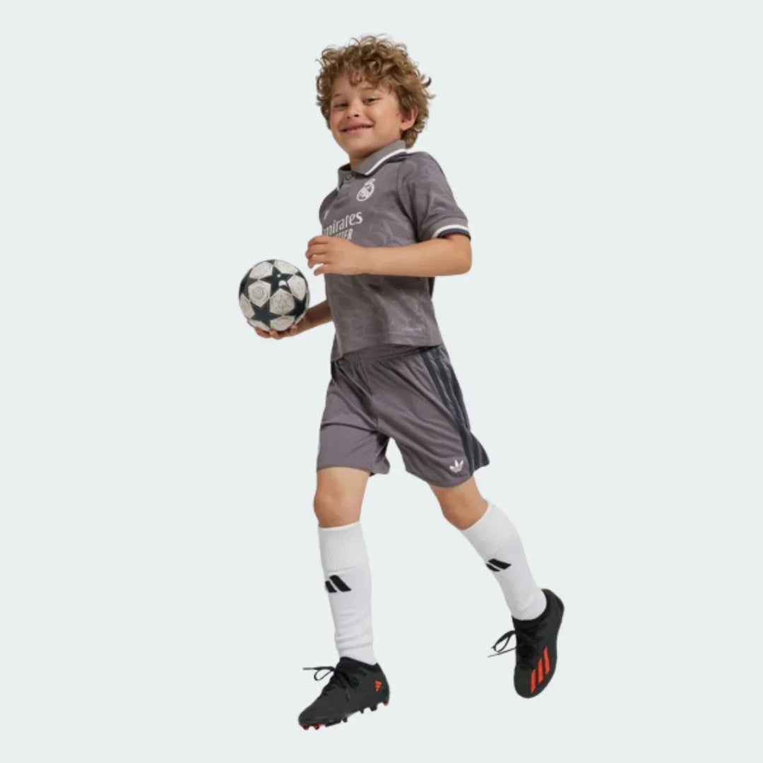 Menino sorridente com kit de futebol infantil Real Madrid cinza. Camisa cinza com escudo branco, shorts cinza com listras escuras, meias brancas, segurando bola.