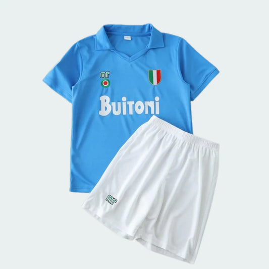 Conjunto infantil de camisa azul claro e shorts branco do Napoli 87/88, com logos NR e Buitoni, em fundo cinza.