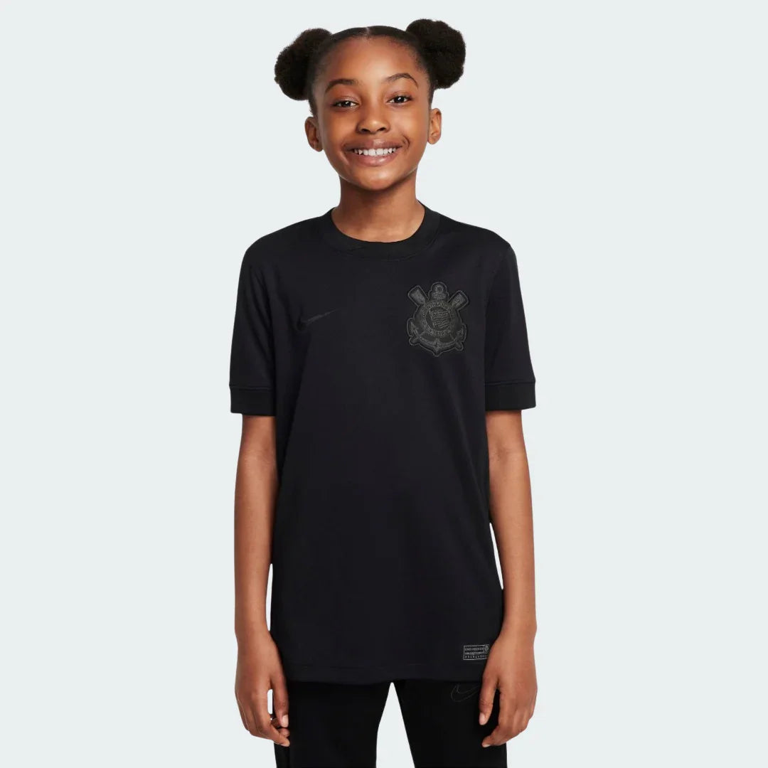 Jovem garota sorrindo vestindo camisa de futebol infantil preta do Timão, com escudo e logo Nike em tom escuro, em fundo branco.