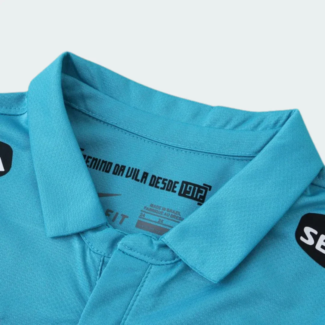 Vista superior do colar de uma camisa esportiva infantil azul-turquesa. Detalhes internos: 'ENINO DA VILA DESDE 1912', logo Nike, 'MADE IN BRAZIL'. Patch 'SE' visível na lateral.