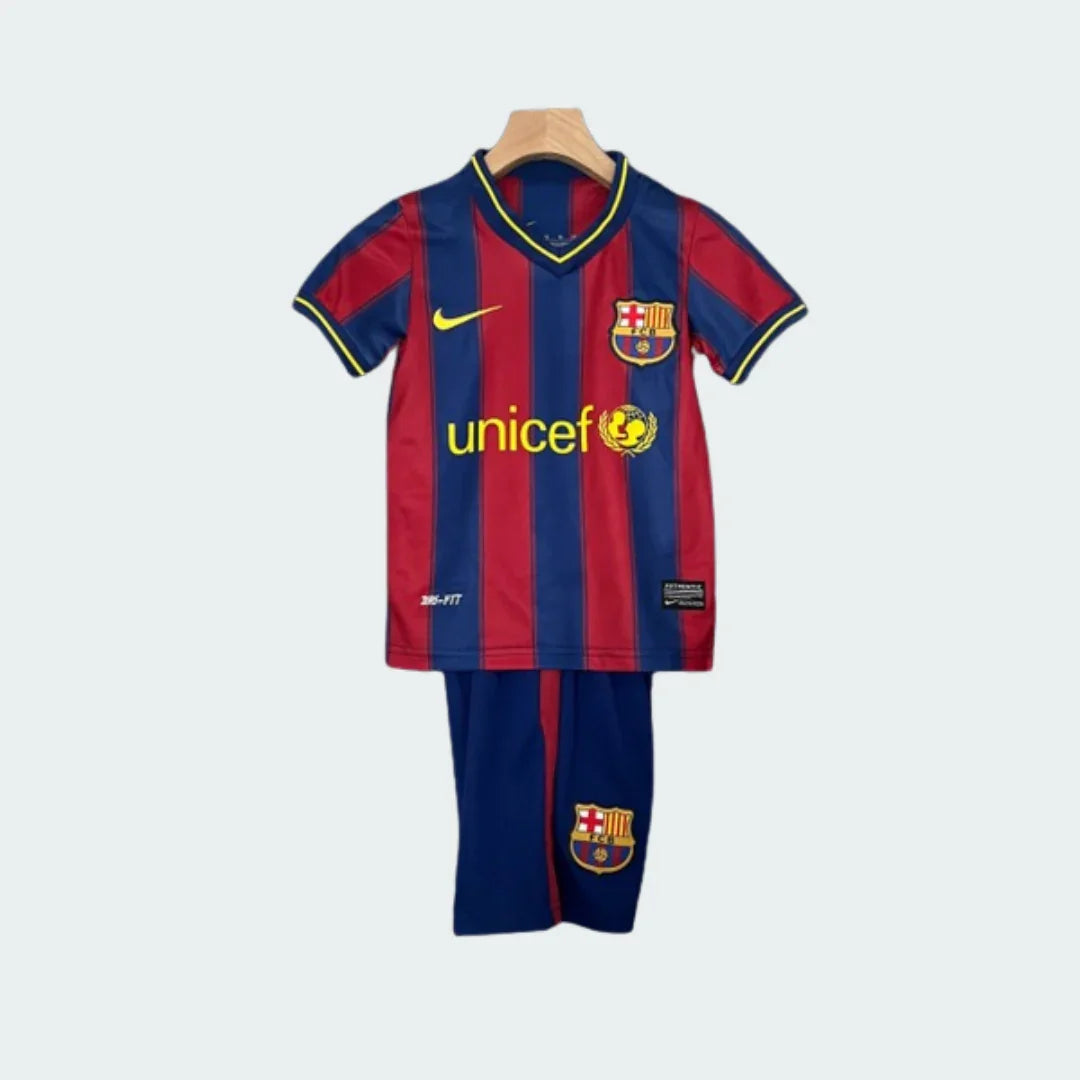 Kit infantil retrô Barcelona com camisa listrada vertical em vermelho e azul, shorts azuis e escudo do time. Pendurado em fundo branco.
