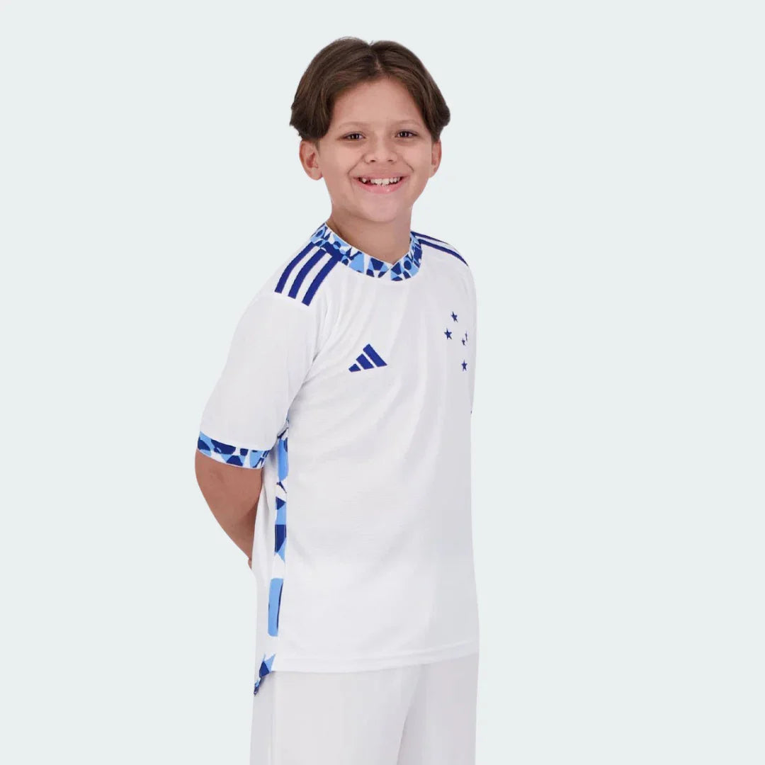 Menino sorridente vestindo camisa de futebol juvenil branca do Cruzeiro, shorts brancos. Detalhes azuis da Adidas, estrelas no peito.