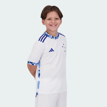 Menino sorridente vestindo camisa de futebol juvenil branca do Cruzeiro, shorts brancos. Detalhes azuis da Adidas, estrelas no peito.