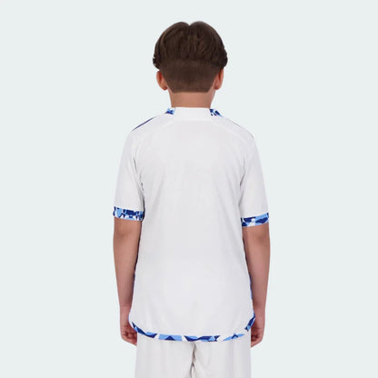 Menino de costas vestindo camisa de futebol branca com padrões geométricos azuis e azul claro nos punhos, gola e barra. Shorts brancos.