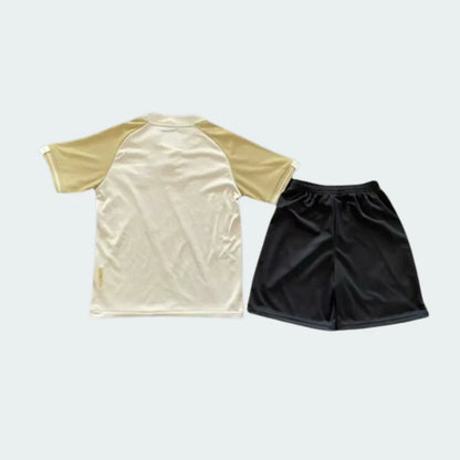 Foto de cima de um kit infantil de futebol com camisa bege e mangas caqui, e shorts esportivos pretos em fundo neutro.