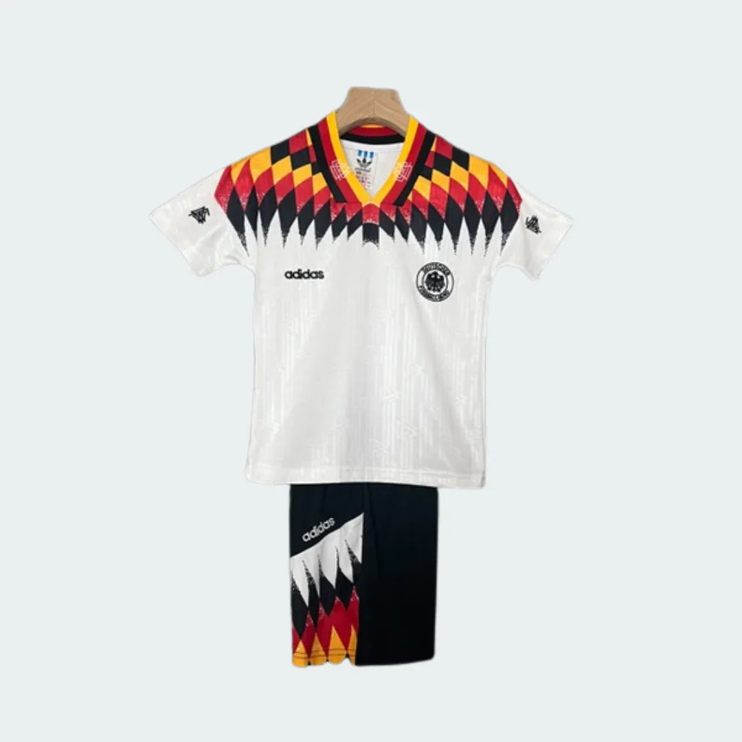 Kit infantil retrô da Alemanha 1994 com camisa branca e shorts pretos. A camisa tem gola V e detalhes em preto, vermelho e amarelo.