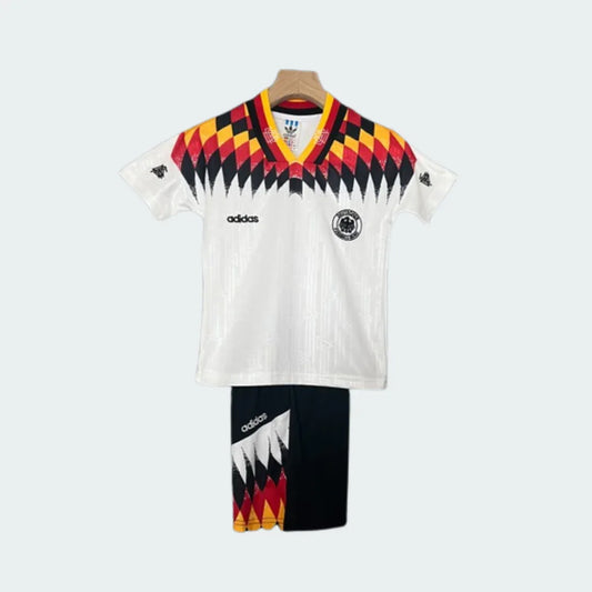 Kit infantil retrô da Alemanha 1994 com camisa branca e shorts pretos. A camisa tem gola V e detalhes em preto, vermelho e amarelo.