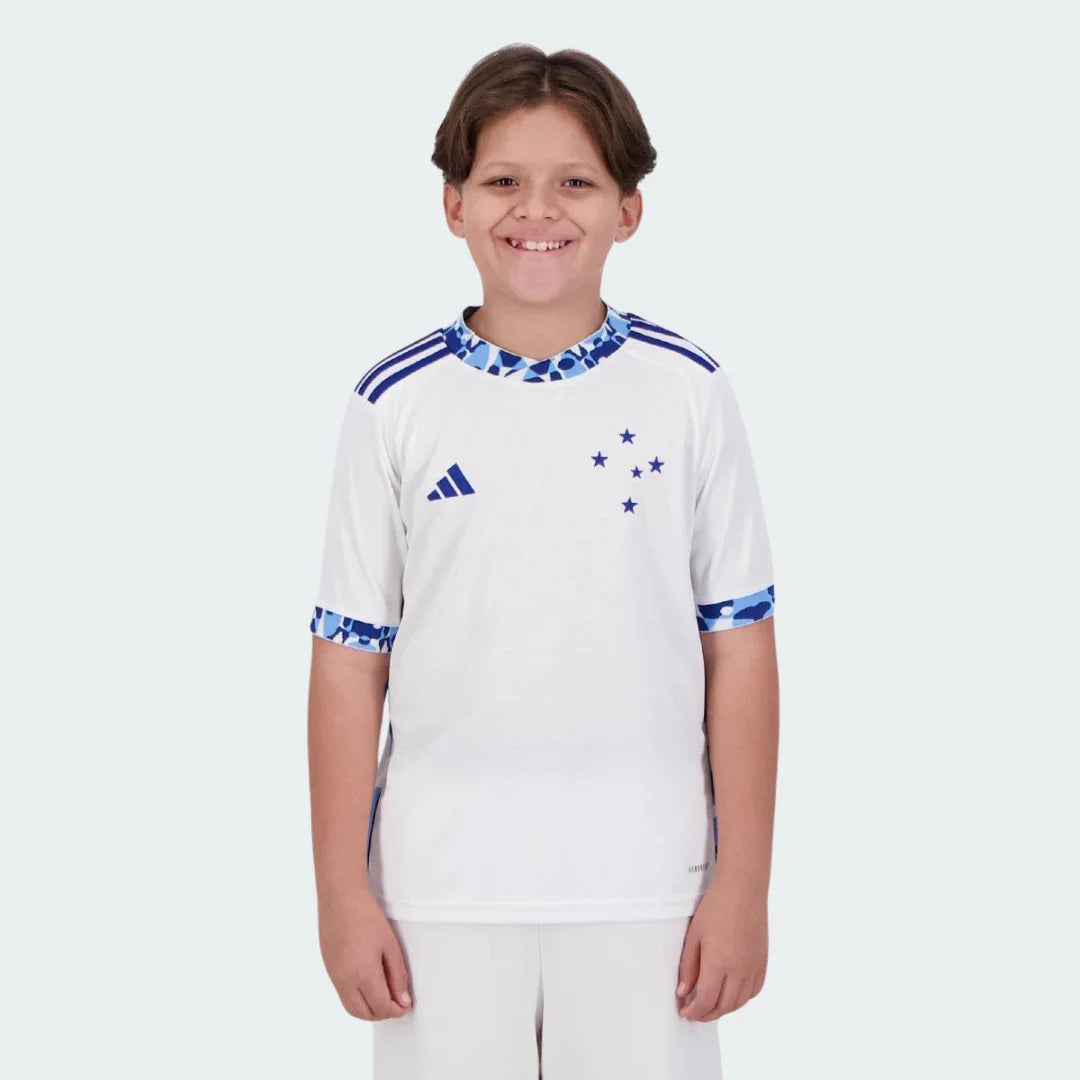 Menino sorridente veste kit infantil branco Cabuloso II 24/25 com detalhes azuis, logo Adidas e cinco estrelas no peito.