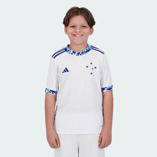 Menino sorridente veste kit infantil branco Cabuloso II 24/25 com detalhes azuis, logo Adidas e cinco estrelas no peito.