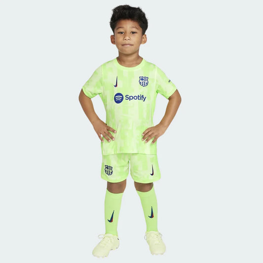 Menino sorridente veste kit de futebol infantil Barcelona III 24/25, camisa e shorts verde limão com patrocínio Spotify e escudo do time.