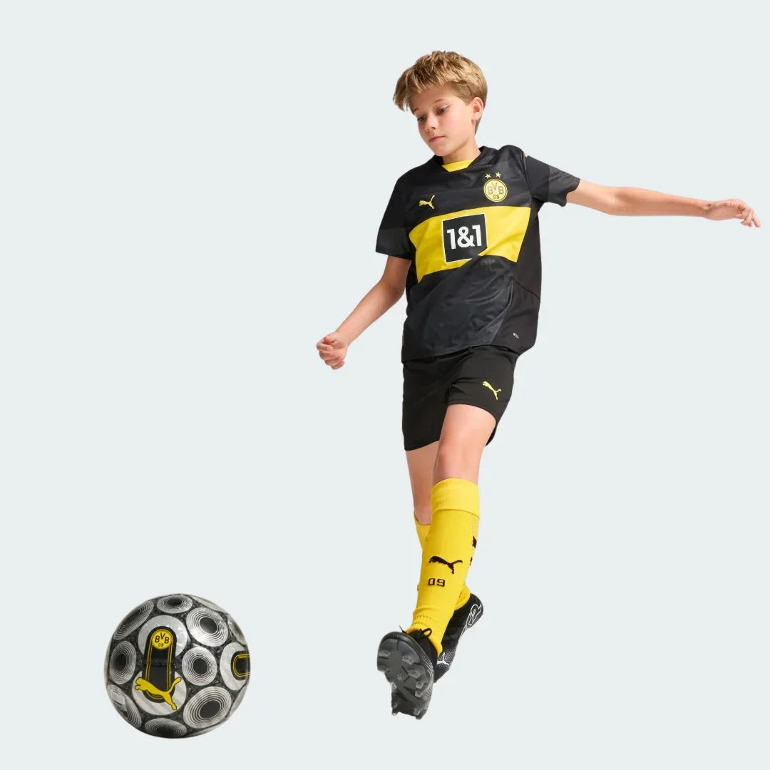 Criança vestindo o kit preto e amarelo do Borussia Dortmund II, chutando uma bola de futebol preta e cinza.