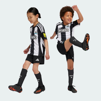 Duas crianças sorrindo vestindo o Kit Infantil Newcastle I 24/25, camisa listrada preta e branca, shorts e meias pretas. Fundo branco.