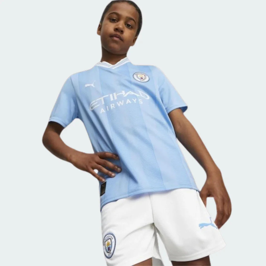 Menino veste o Kit Infantil Manchester City I 23/24, camisa azul clara com listras, shorts brancos e escudo do time.