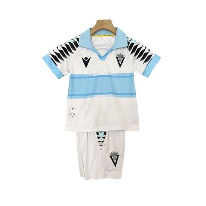 Kit infantil do Cádiz 24/25 (camisa e shorts) em branco com listras azuis claras, escudos do clube e Macron, pendurado.