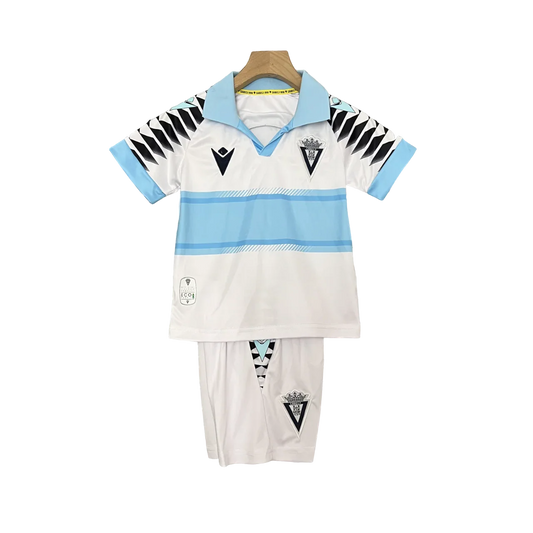 Kit infantil do Cádiz 24/25 (camisa e shorts) em branco com listras azuis claras, escudos do clube e Macron, pendurado.