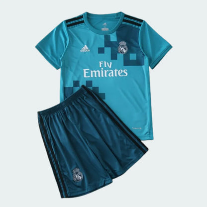 Kit infantil Real Madrid azul-claro com detalhes pixelados, logo Adidas, escudo e Fly Emirates. Shorts azul-escuro com listras e escudo.