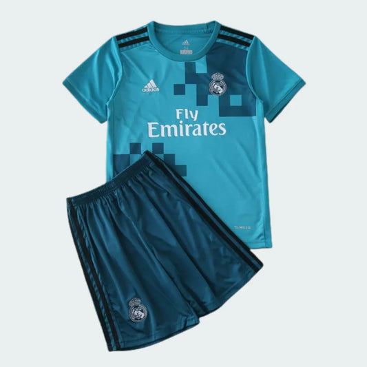 Kit infantil Real Madrid azul-claro com detalhes pixelados, logo Adidas, escudo e Fly Emirates. Shorts azul-escuro com listras e escudo.