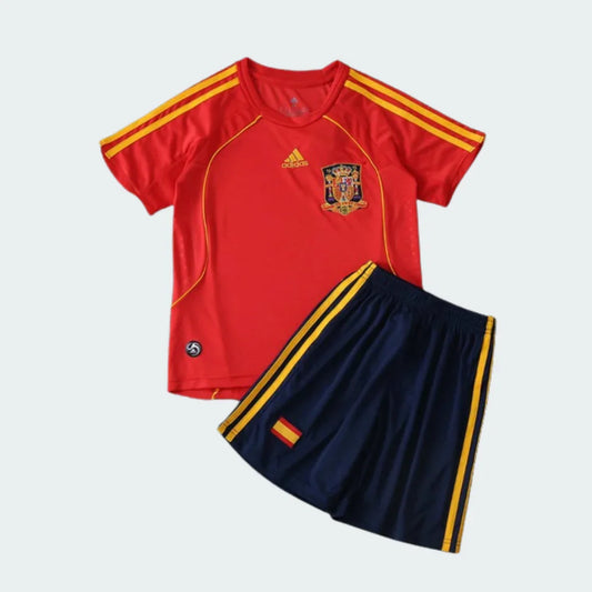 Kit infantil retrô da Espanha com camisa vermelha, listras amarelas e escudo, e shorts azul-marinho com listras amarelas e bandeira.