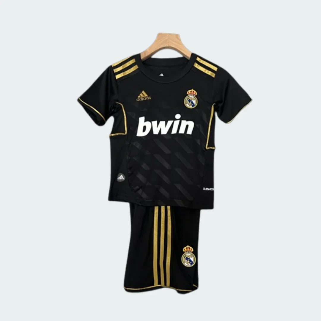 Kit de futebol infantil do Real Madrid 11/12, camisa e shorts pretos com detalhes e listras douradas, logo Adidas e escudo do time. Sponsor bwin branco.